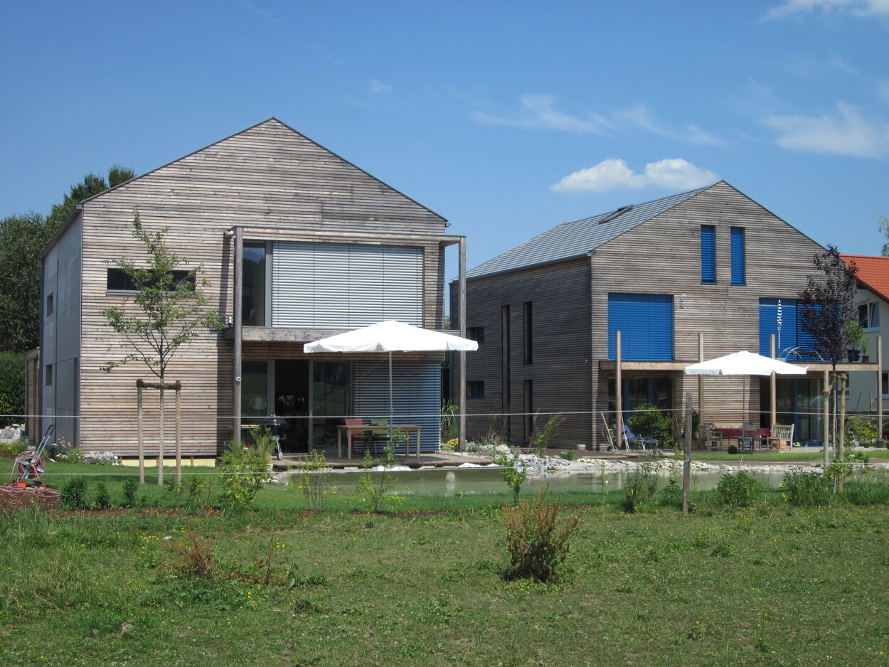 Zwei moderne Passivhäuser mit Holzfassade stehen in Thalham unter einem klaren blauen Himmel in einer grünen Landschaft.