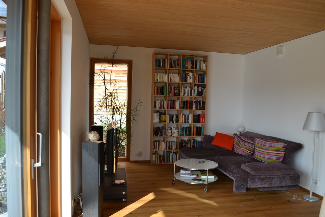 Ein gemütliches Wohnzimmer im Passivhaus Thalham mit Sofa, Bücherregal und großem Fenster, das viel Licht hereinlässt.