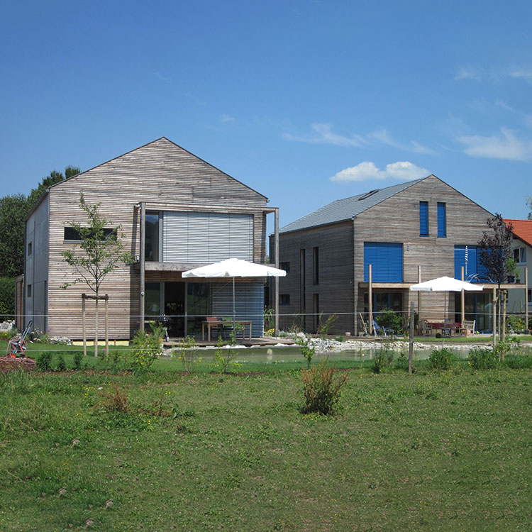 Zwei moderne Passivhäuser in Thalham stehen nebeneinander mit Sonnenschirmen im Garten unter blauem Himmel.