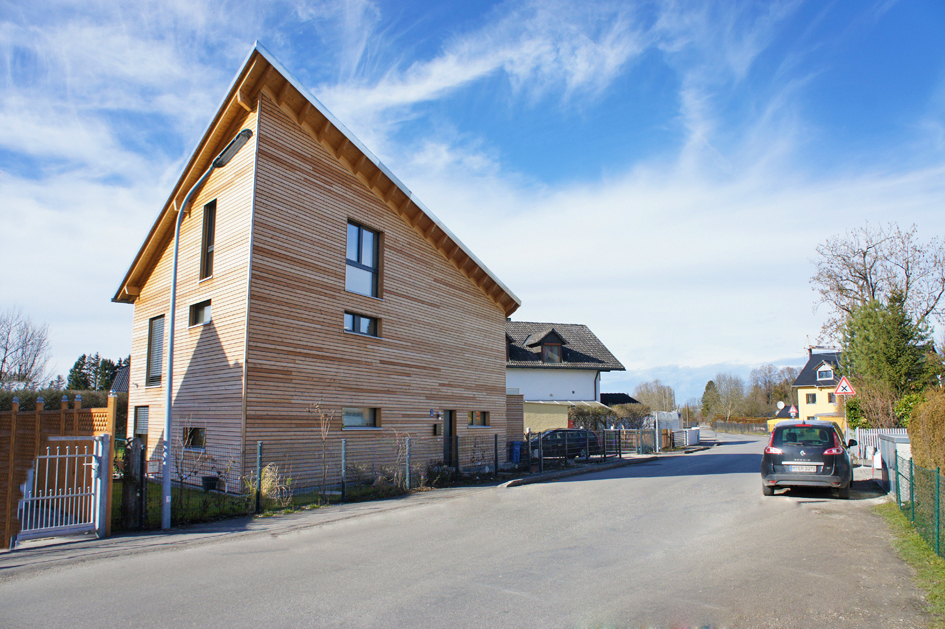 Ein modernes Passivhaus mit Holzfassade steht in München an einer ruhigen Straße unter strahlend blauem Himmel.
