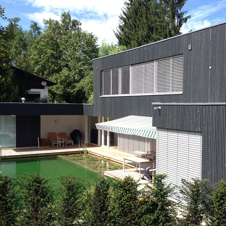Ein modernes Passivhaus in Brannenburg mit Holzverkleidung und einem natürlichen Schwimmteich im sonnigen Garten.
