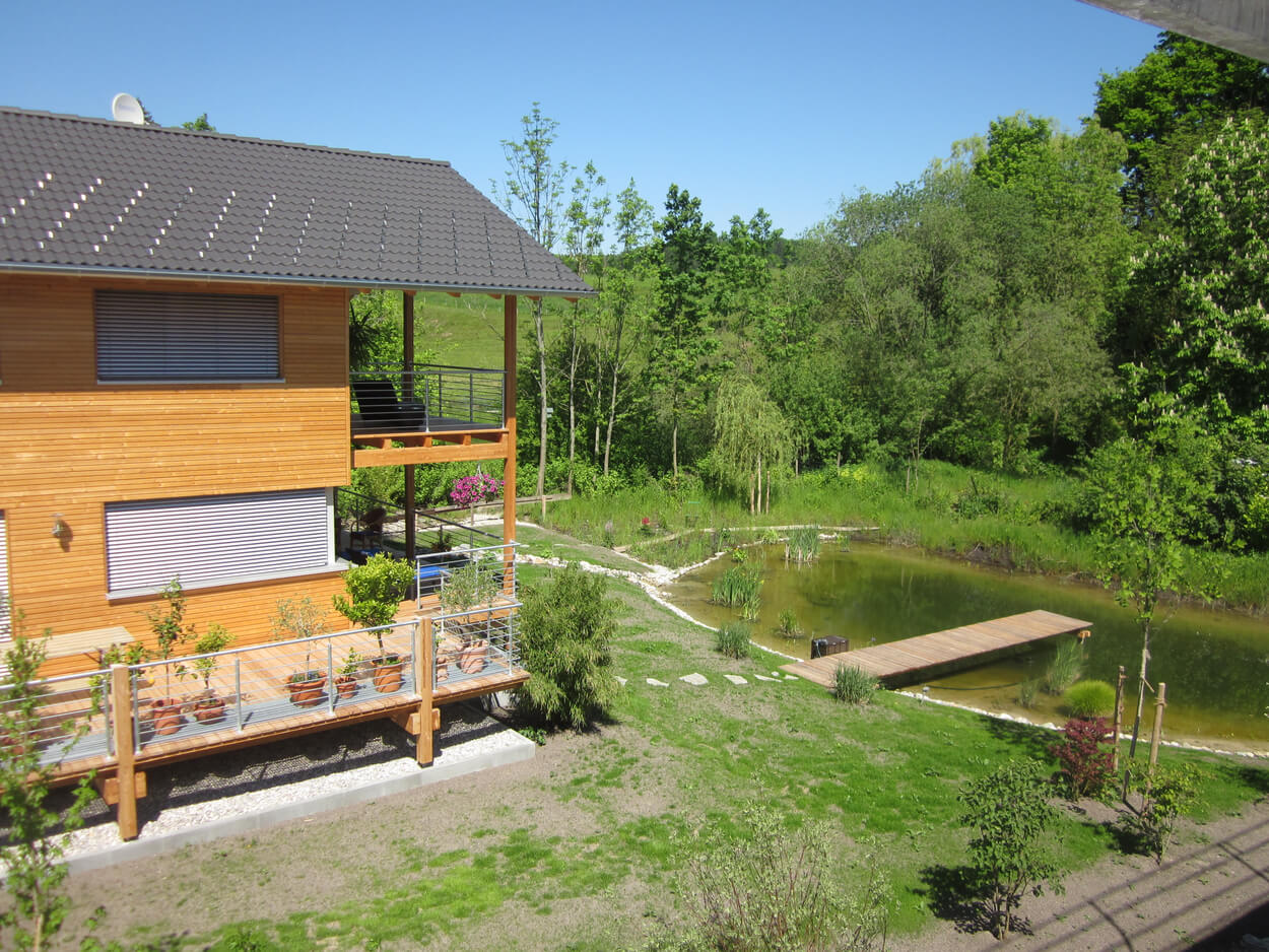 Ein modernes Holz-Passivhaus mit Terrasse steht idyllisch neben einem Teich in der grünen Landschaft von Glonn.