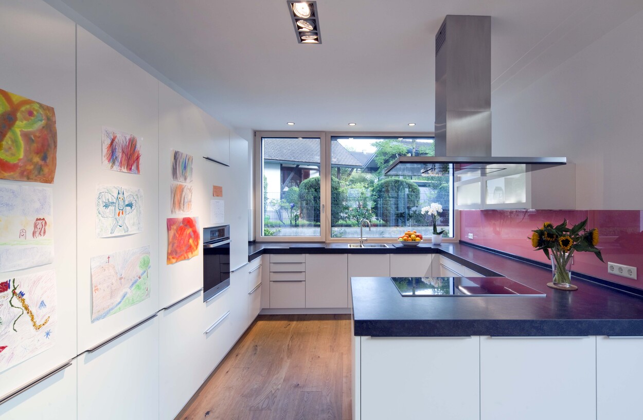 Moderne Küche in einem Passivhaus mit Kinderzeichnungen an der Wand und Blick auf den Garten durch ein großes Fenster.