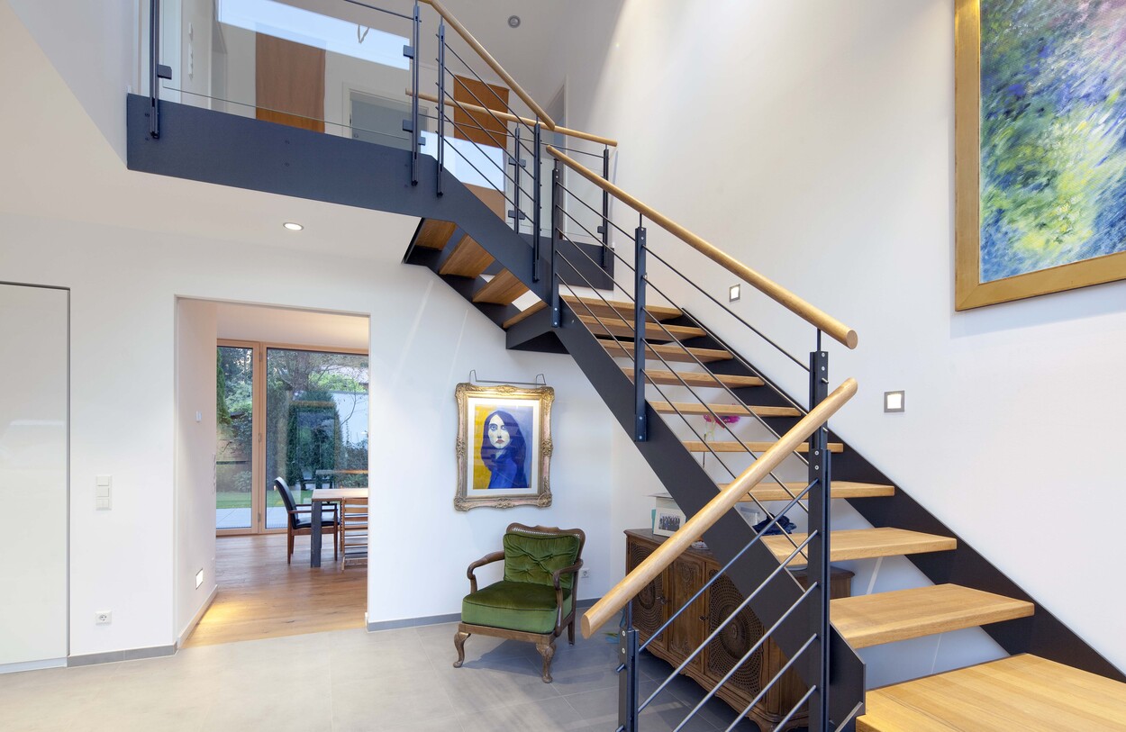 Eine moderne Holztreppe im Passivhaus in Ismaning führt an Kunstwerken und einem eleganten Sessel vorbei zur Galerie.