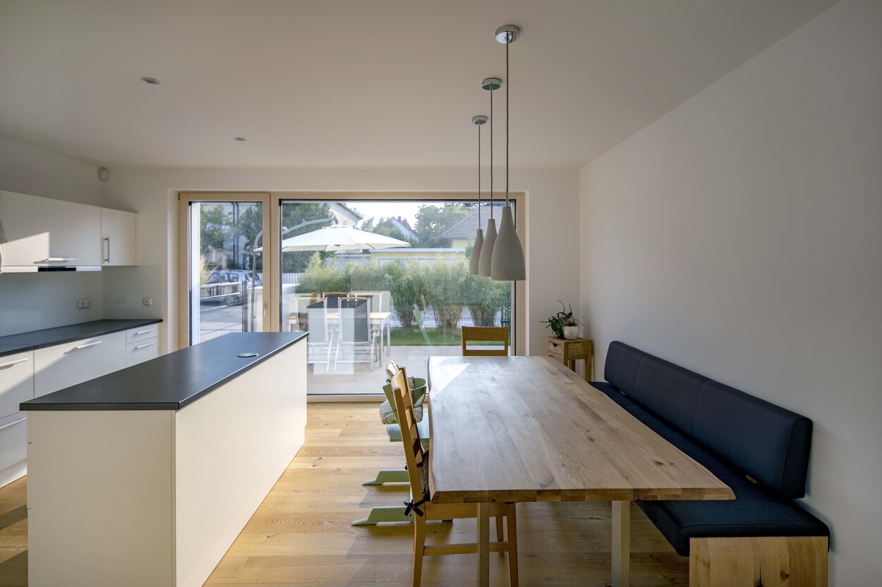 Moderne Küche in einem Münchner Doppelhaus mit großem Holztisch und Blick auf den grünen Garten durch eine Glasfront.