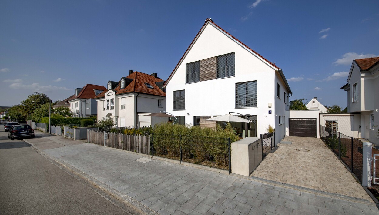 Ein modernes Doppelhaus mit weißer Fassade steht unter blauem Himmel in einer ruhigen Wohnstraße in München.