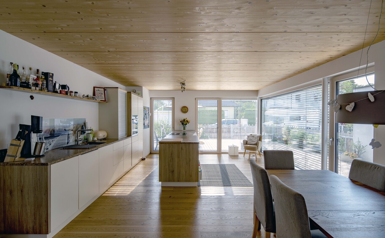 Eine moderne Küche in einem Münchner Doppelhaus mit Holzdecke und großen Fenstern, die viel Licht hereinlassen.