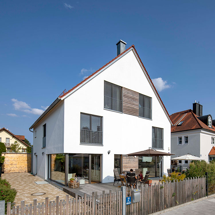 Ein modernes Doppelhaus in München mit Garten und Terrasse, umgeben von einem Holzzaun, steht unter klarem blauem Himmel.
