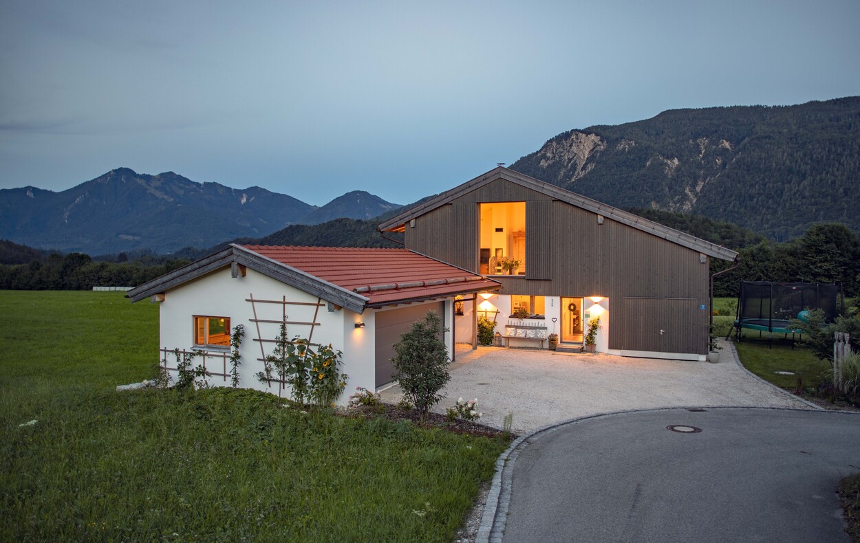 Ein beleuchtetes Holzhaus in Schleching steht stimmungsvoll vor einer beeindruckenden Bergkulisse in der Dämmerung.
