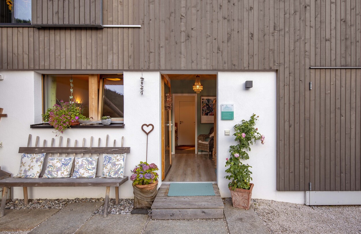 Der Eingang des Holzhauses in Schleching ist mit blühenden Pflanzen und einer rustikalen Bank einladend dekoriert.