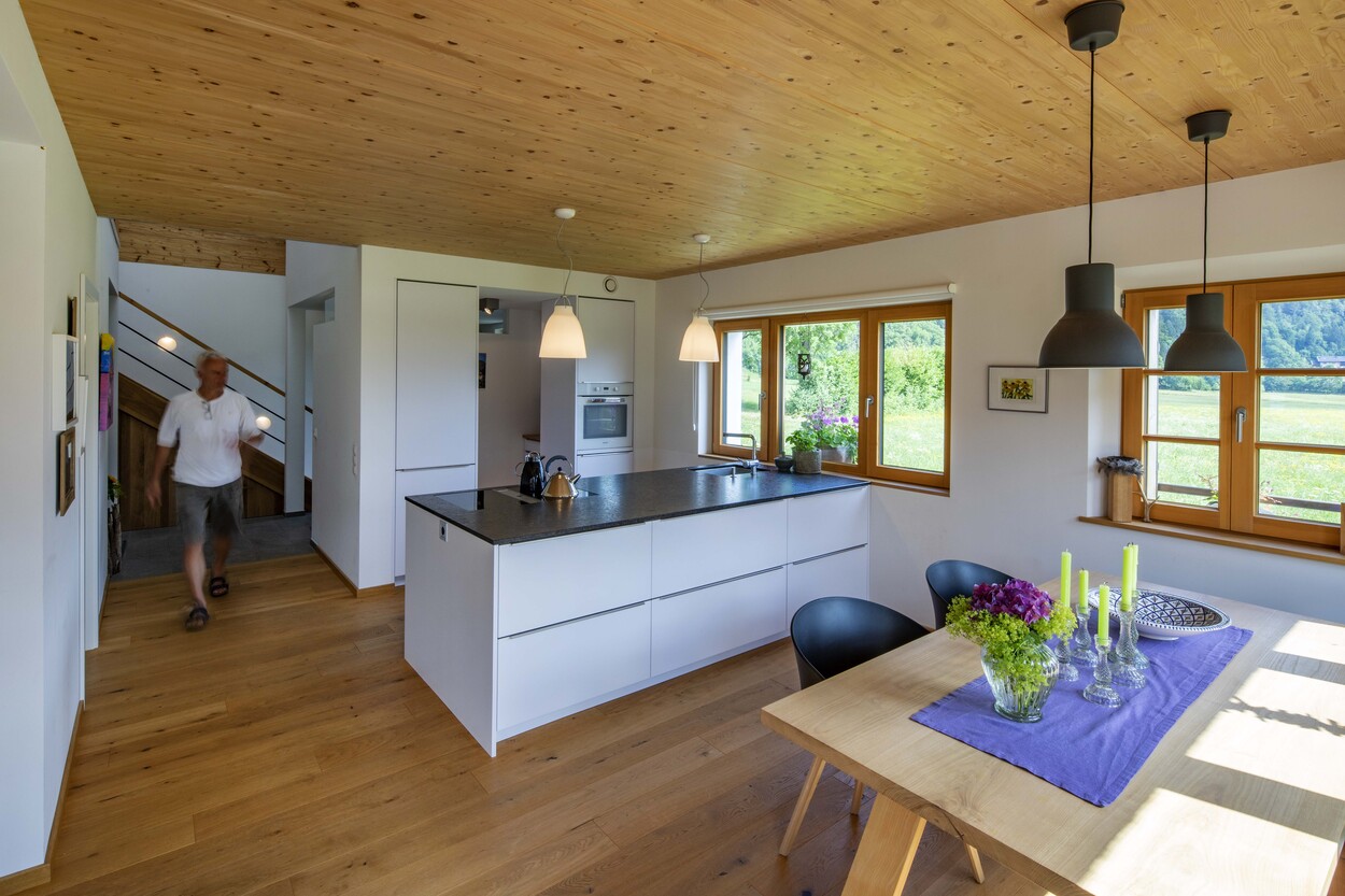 In einem modernen Holzhaus in Schleching betritt ein Mann die helle Küche mit Esstisch und großen Fenstern.