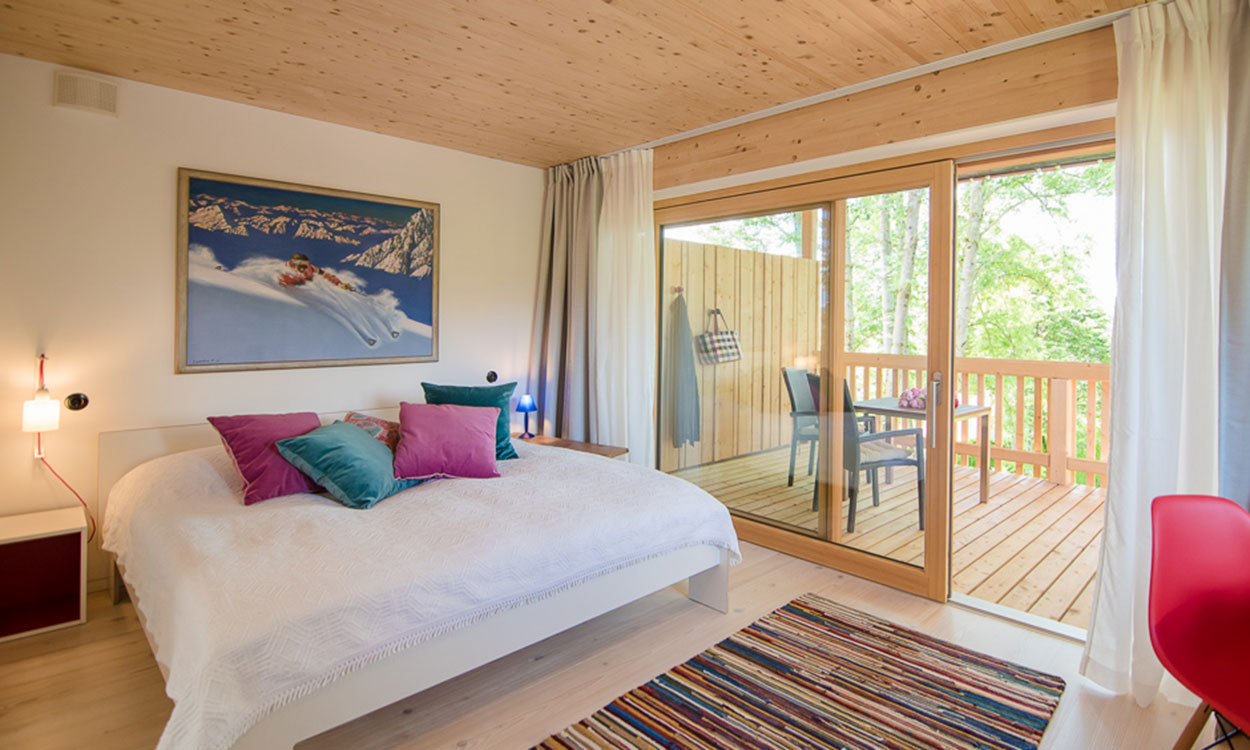 Ein gemütliches Schlafzimmer im Passivhaus mit Holzdecke, bunter Bettwäsche und Zugang zu einem Balkon mit Blick ins Grüne.