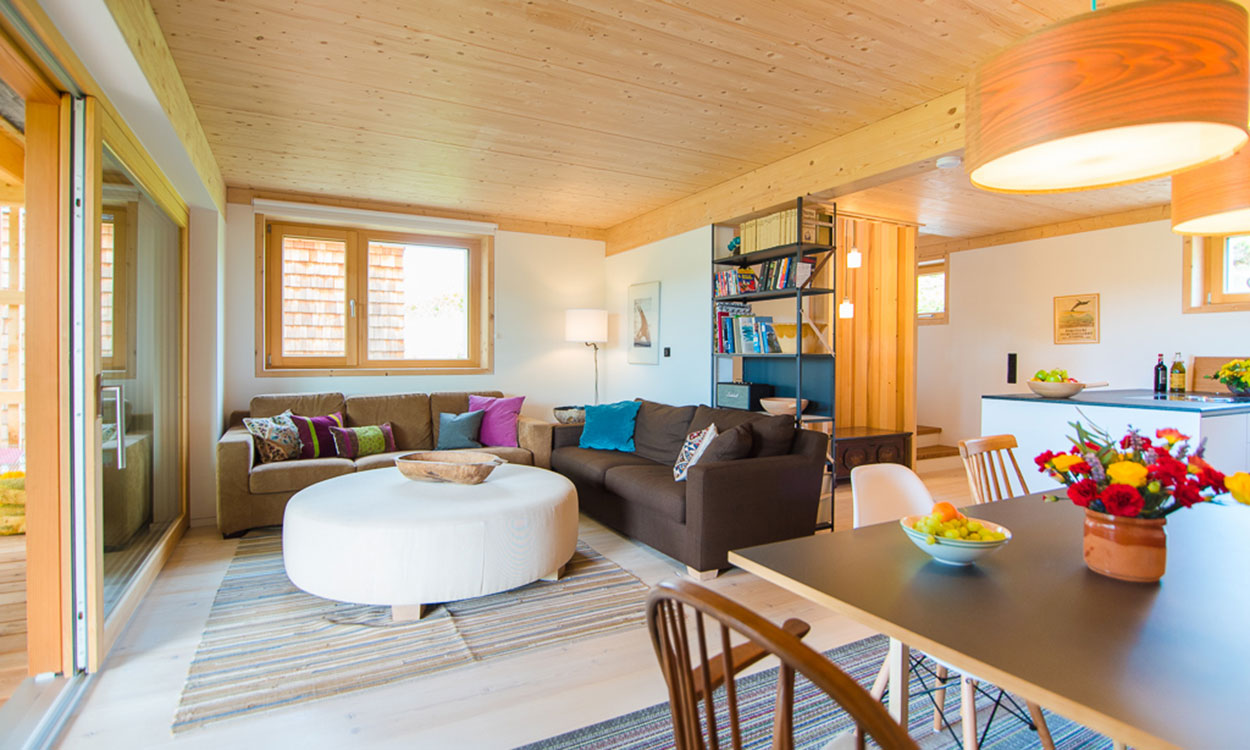 Ein gemütliches Wohnzimmer im Passivhaus mit Holzmöbeln, bunten Kissen und einem großen Tisch lädt zur Entspannung ein.