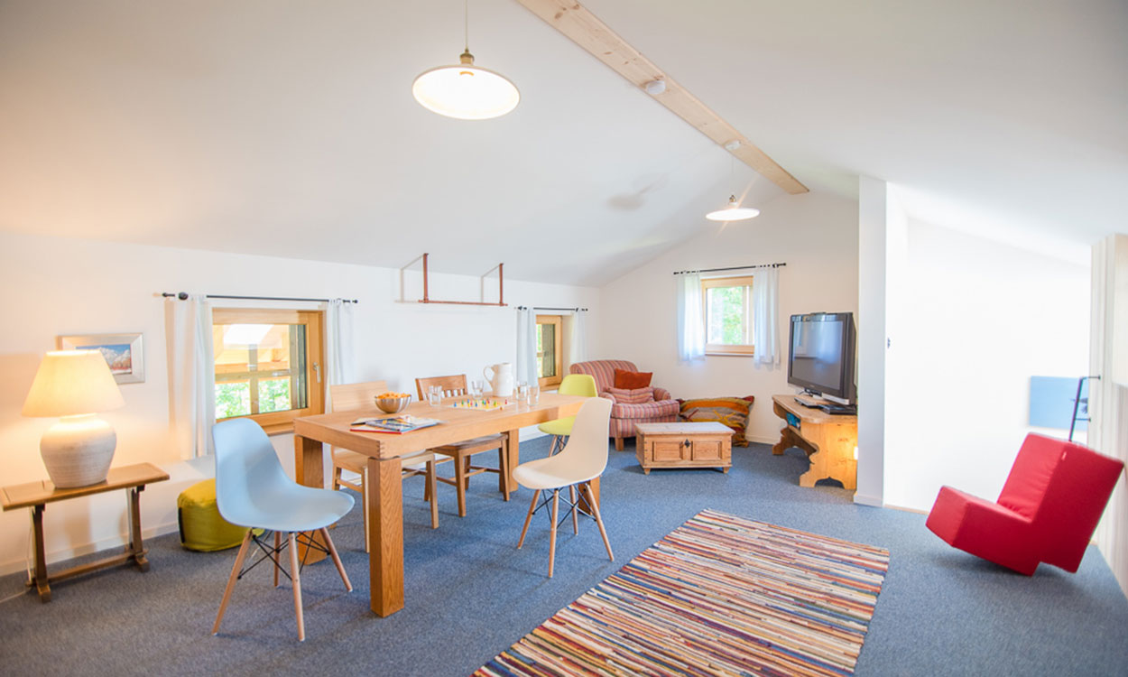 Ein helles Wohnzimmer im Passivhaus mit Holztisch, buntem Teppich und gemütlicher Sitzecke vor großen Fenstern.