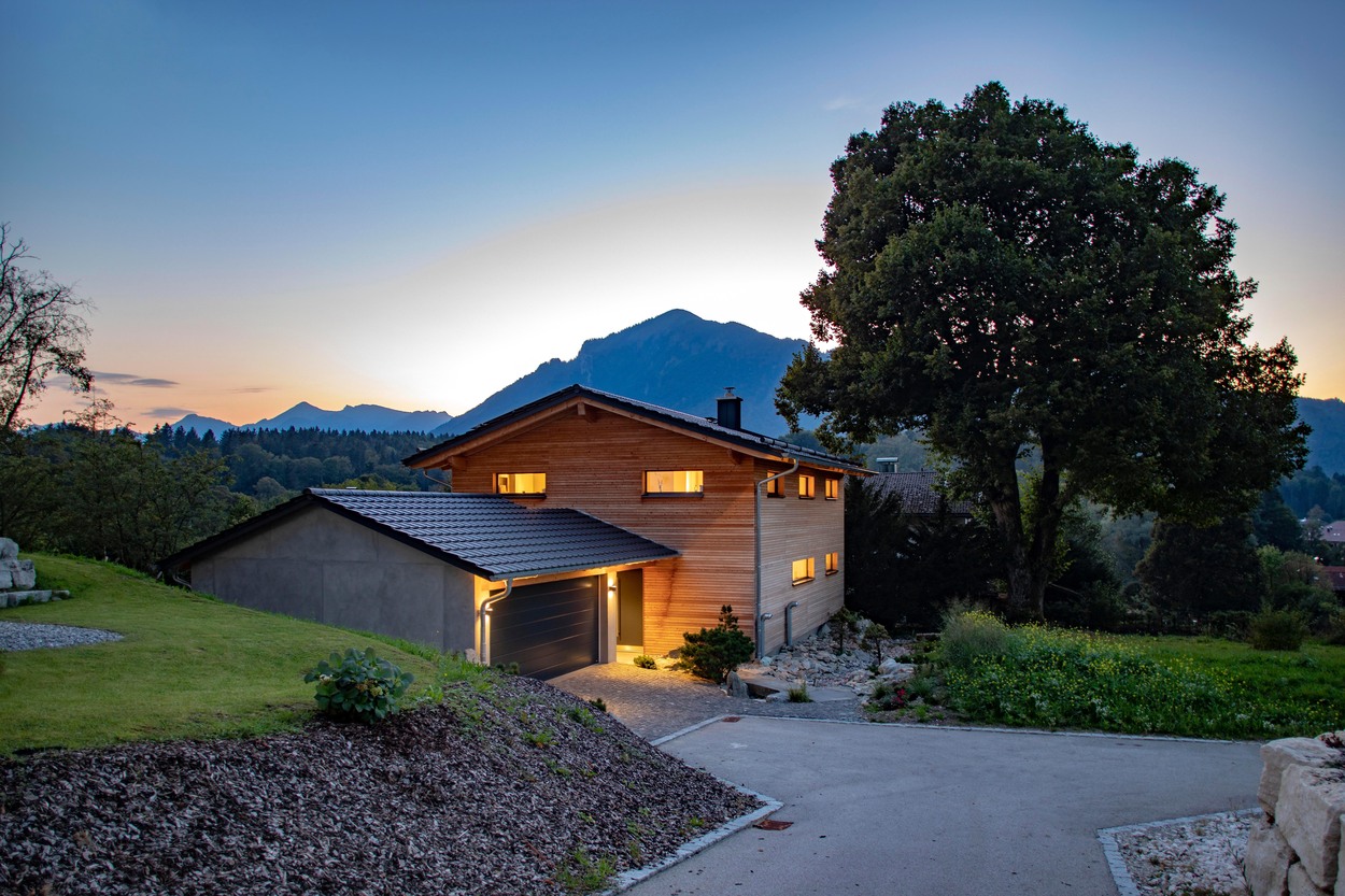 Ein beleuchtetes Holzhaus in Marquartstein steht malerisch vor einem Bergpanorama im Chiemgau bei Sonnenuntergang.