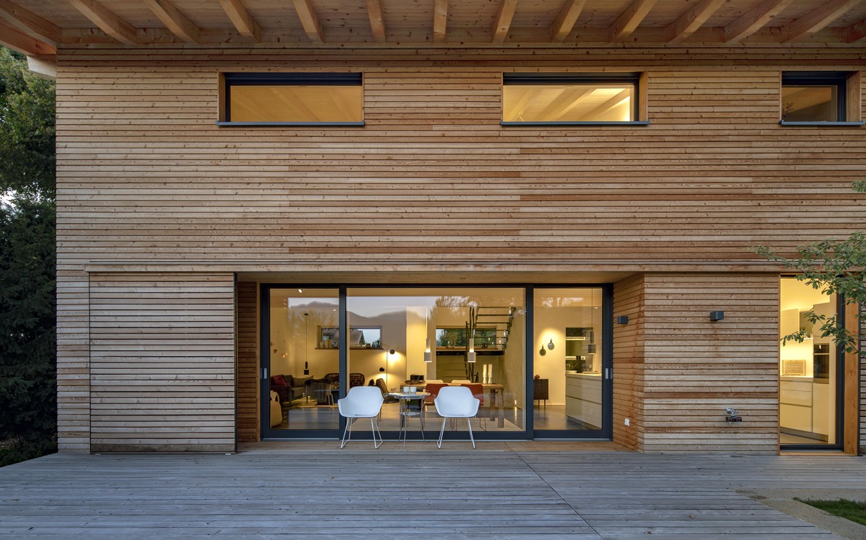 Ein modernes Holzhaus in Marquartstein mit großen Glasfronten und zwei weißen Stühlen lädt auf der Terrasse zum Verweilen ein.