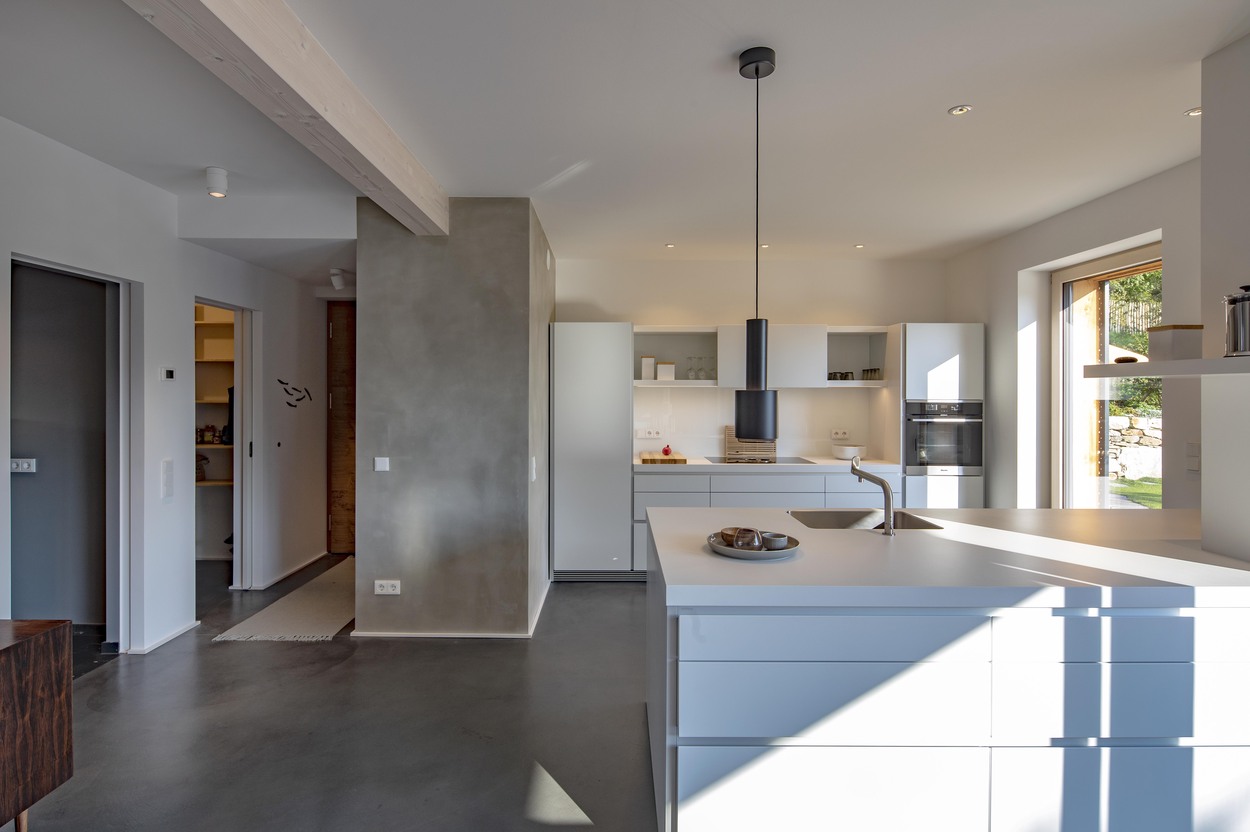 Moderne Küche im Holzhaus in Marquartstein mit weißen Möbeln und großen Fenstern, die viel Licht in den Raum lassen.