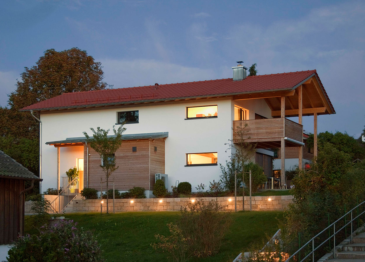 Ein modernes Passivhaus in Oberhaching mit beleuchteter Fassade und Balkon erstrahlt bei Abenddämmerung im gepflegten Garten.