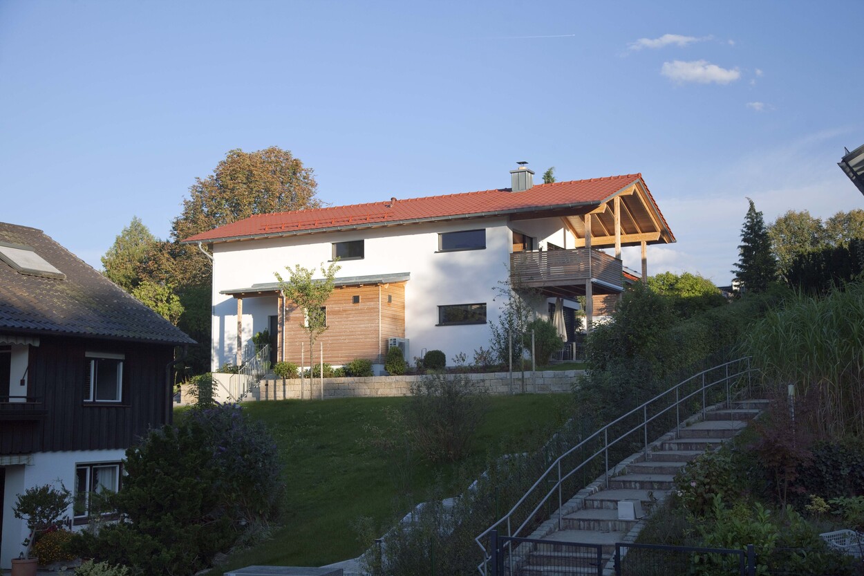 Ein modernes Passivhaus mit rotem Dach steht in Oberhaching, umgeben von einem grünen Garten unter blauem Himmel.