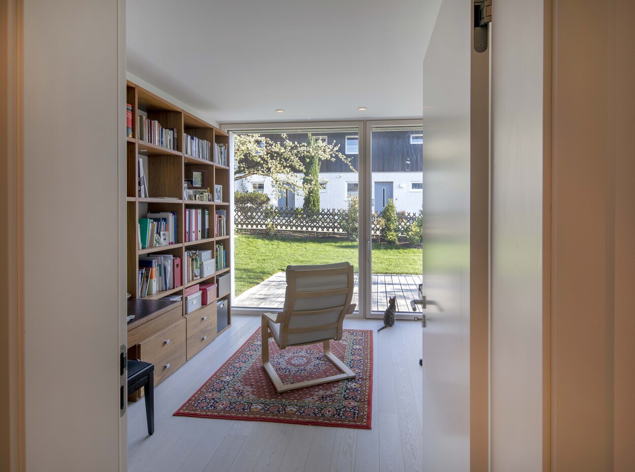 Ein lichtdurchflutetes Arbeitszimmer im Passivhaus Andechs mit einem gemütlichen Sessel und Bücherregal auf einem Teppich.