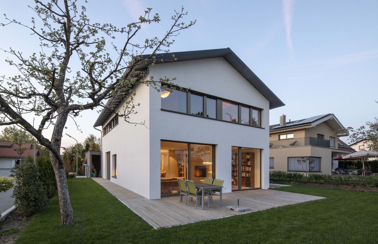 Ein modernes Passivhaus in Andechs mit Holzterrasse und Garten wird von einem blühenden Baum im Vordergrund ergänzt.