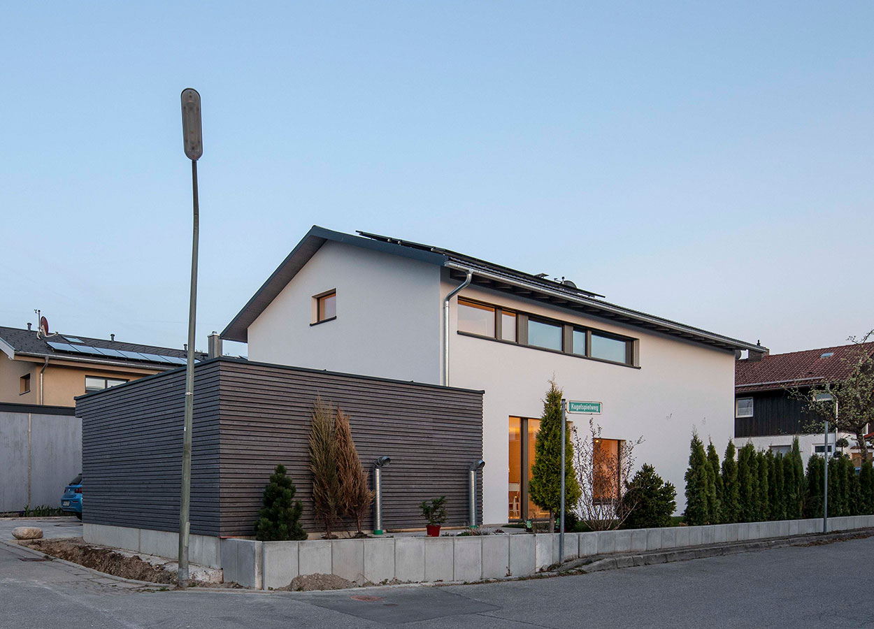 Ein modernes Passivhaus mit Garage in Andechs steht an einer ruhigen Straße, umgeben von Bäumen und klarem Himmel.