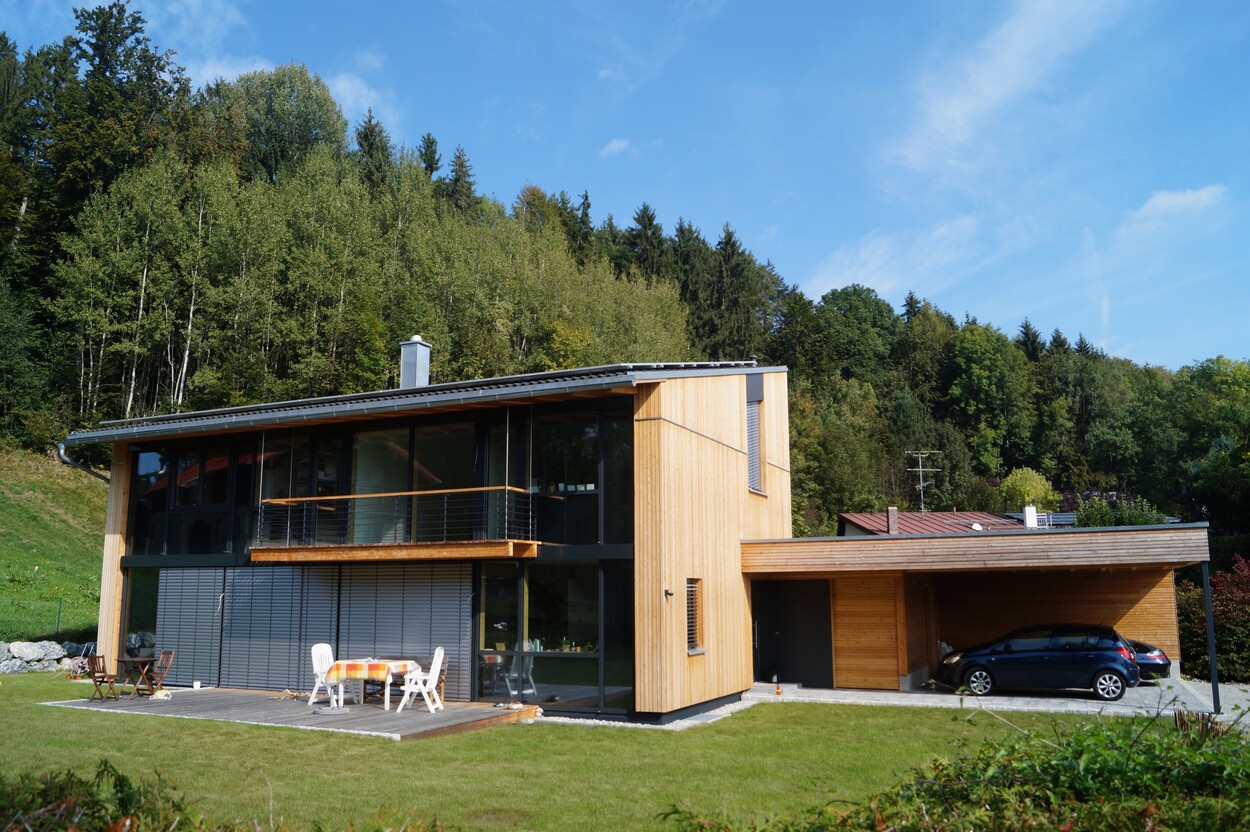 Ein modernes Holz-Einfamilienhaus in Feldkirchen steht vor einem dichten Wald, mit Terrasse und Auto im Carport.