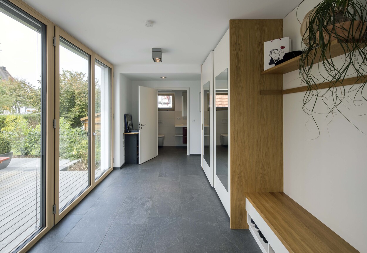 Moderner Flur in einem Passivhaus in München mit großen Fenstern und einem Holzregal voller Pflanzen.