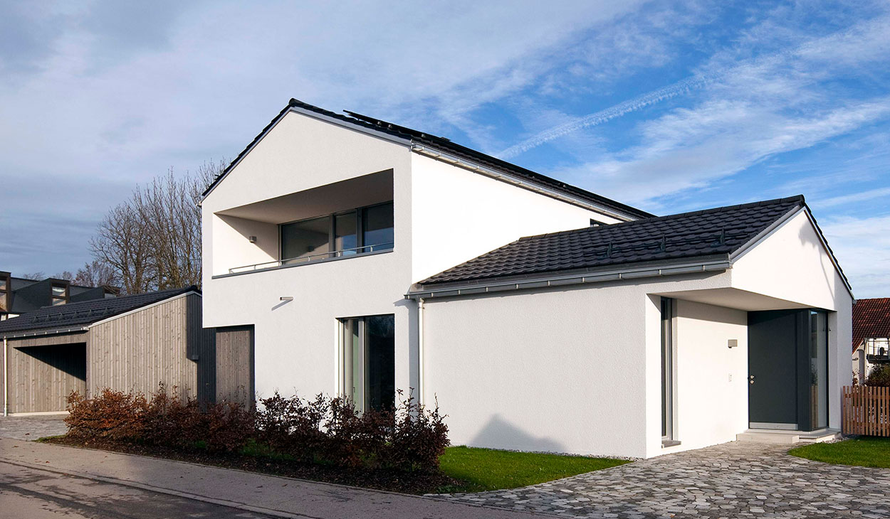 Ein modernes Passivhaus in Bruckmühl mit weißer Fassade und großen Fenstern strahlt unter einem klaren blauen Himmel.