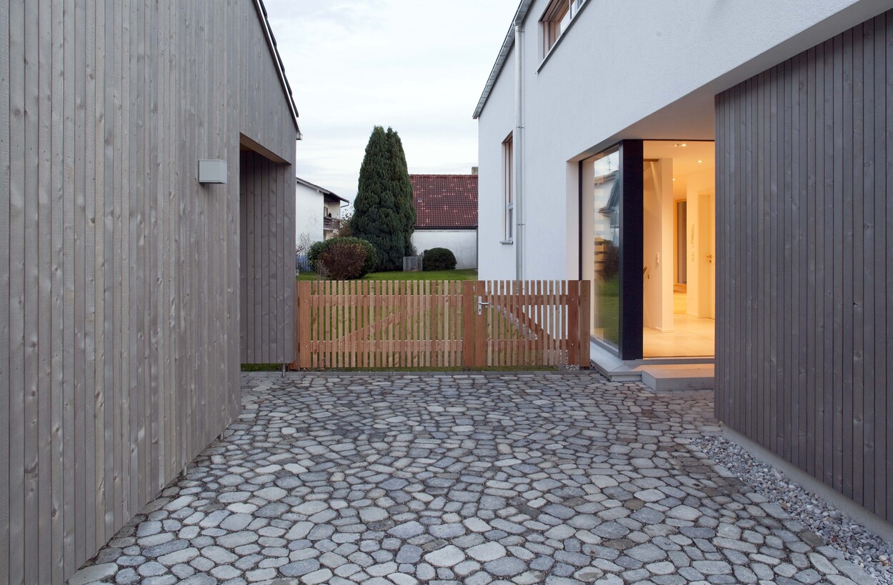 Ein modernes Passivhaus in Bruckmühl zeigt einen gepflasterten Hof mit Holzzaun und großen Fenstern, die viel Licht hereinlassen.