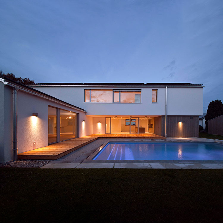 Ein modernes Passivhaus in Bruckmühl erstrahlt bei Abenddämmerung mit beleuchtetem Pool und großen Fenstern.