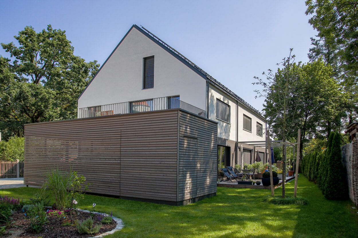 Ein modernes Doppelhaus im Passivhaus-Design steht in einem gepflegten Garten unter klarem Münchener Himmel.