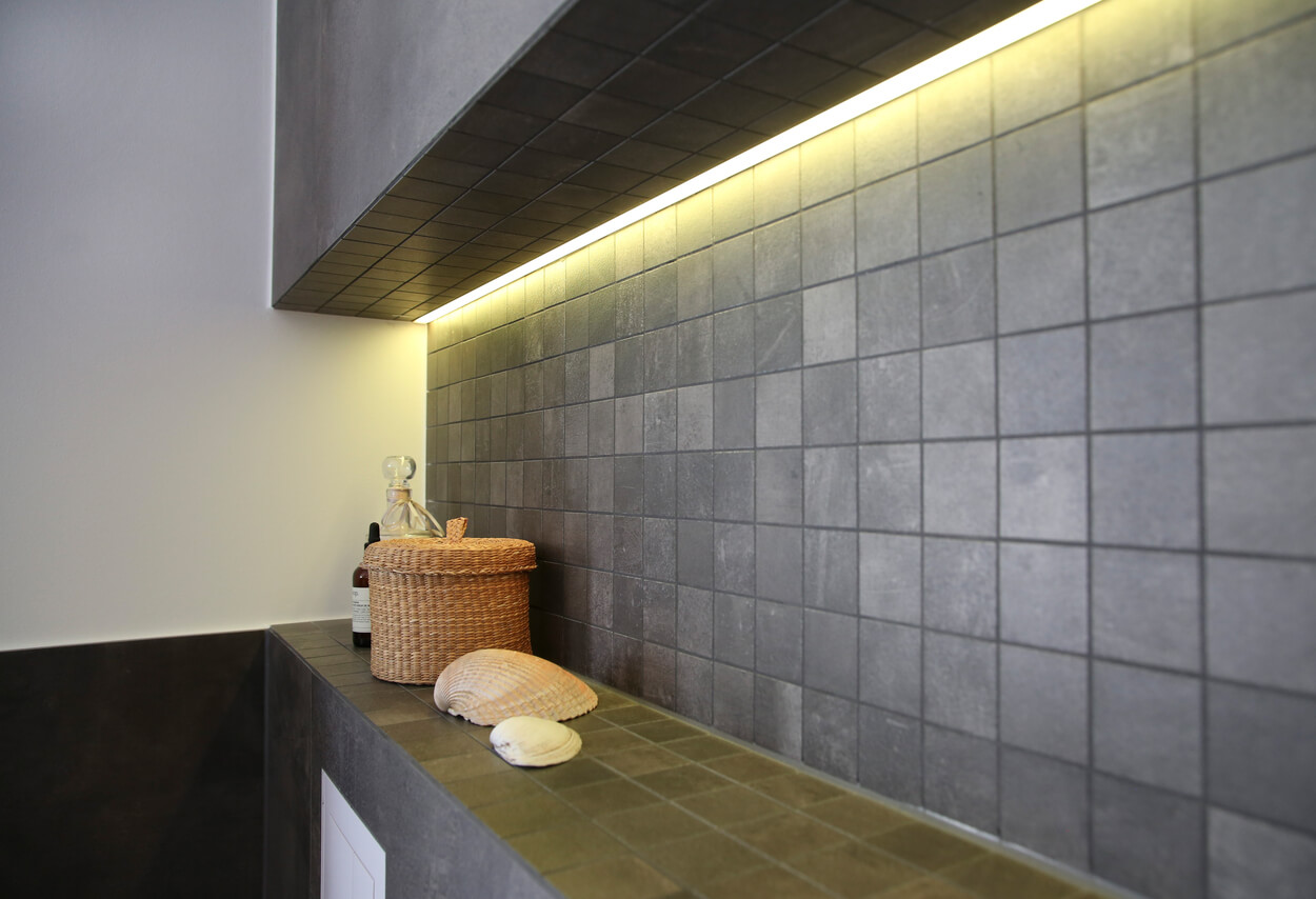 In einem modernen Badezimmer in München sind graue Kacheln, eine geflochtene Dose und Muscheldekorationen stilvoll arrangiert.