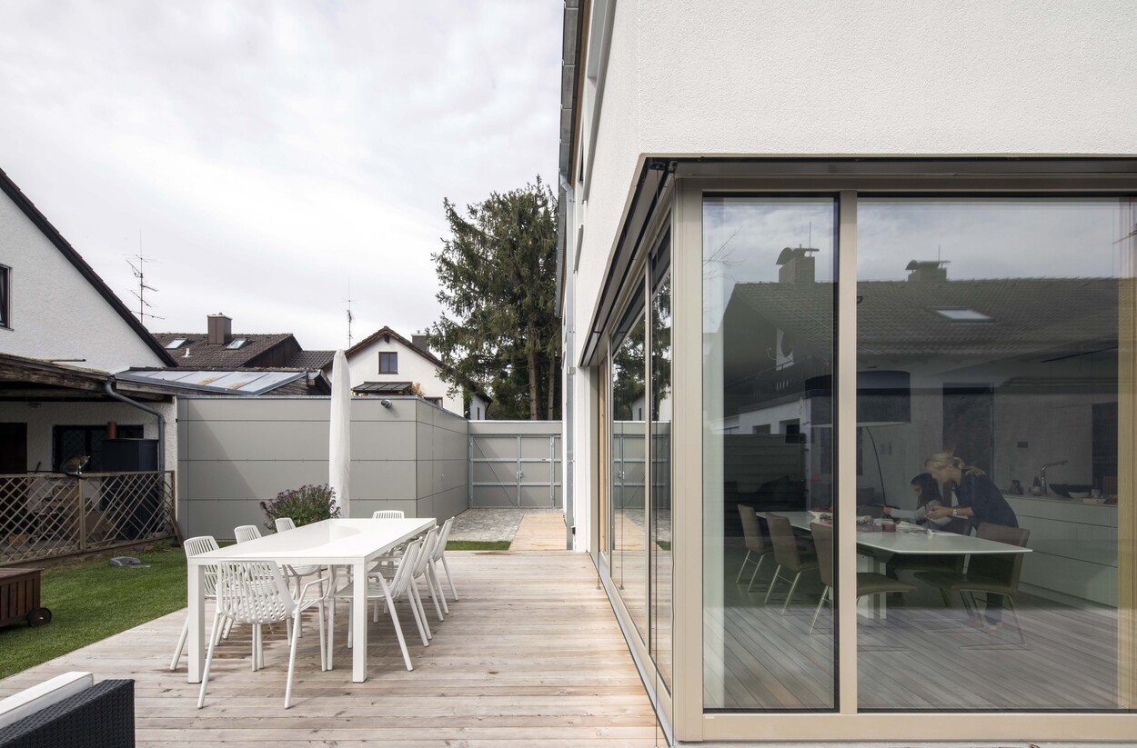 Eine moderne Terrasse in Putzbrunn zeigt einen großen Esstisch neben der Glasfront eines Energiesparhauses in Holzbauweise.