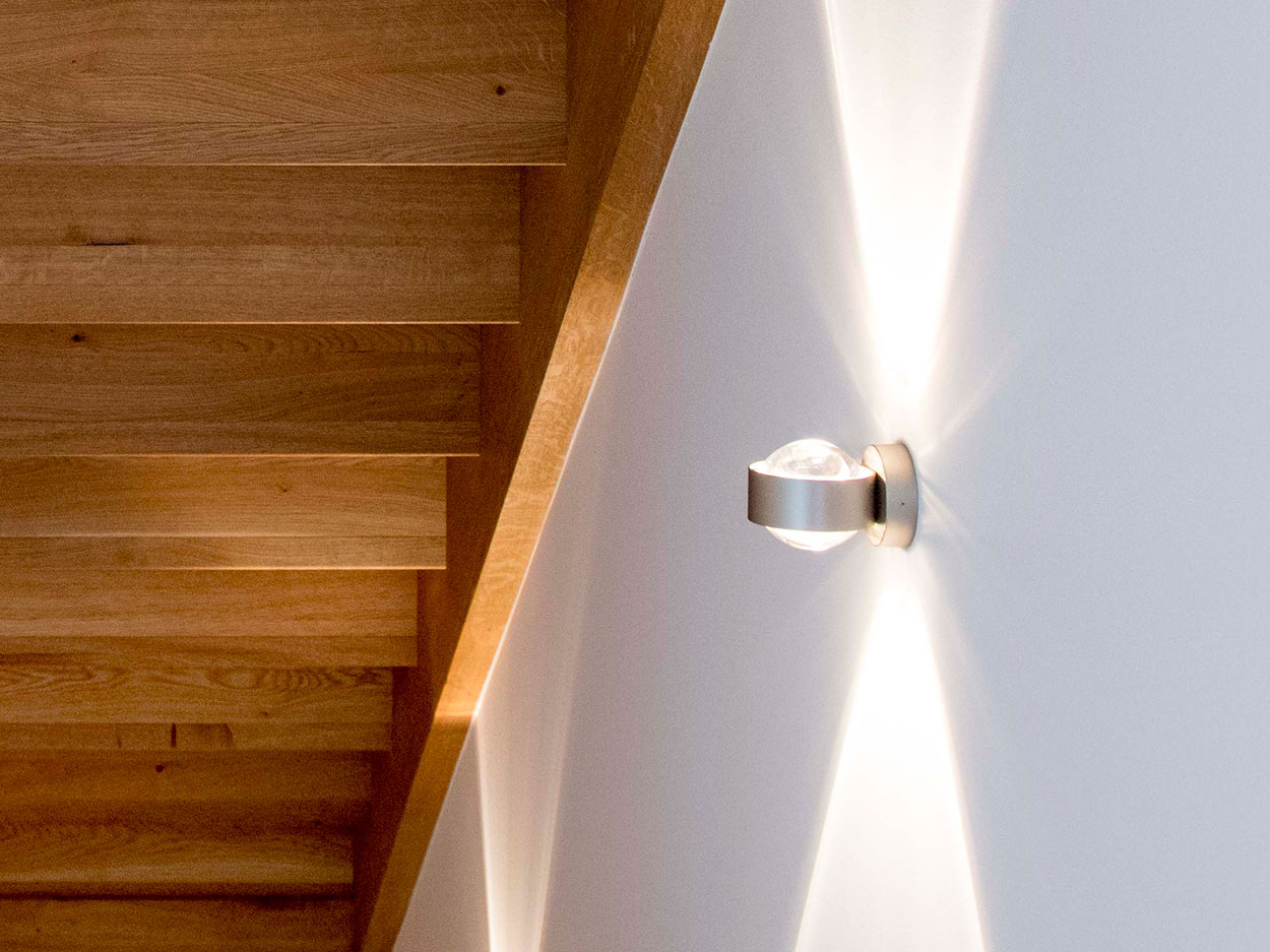 Holztreppenstufen eines Putzbrunner Energiesparhauses werden durch eine moderne Wandleuchte stilvoll beleuchtet.
