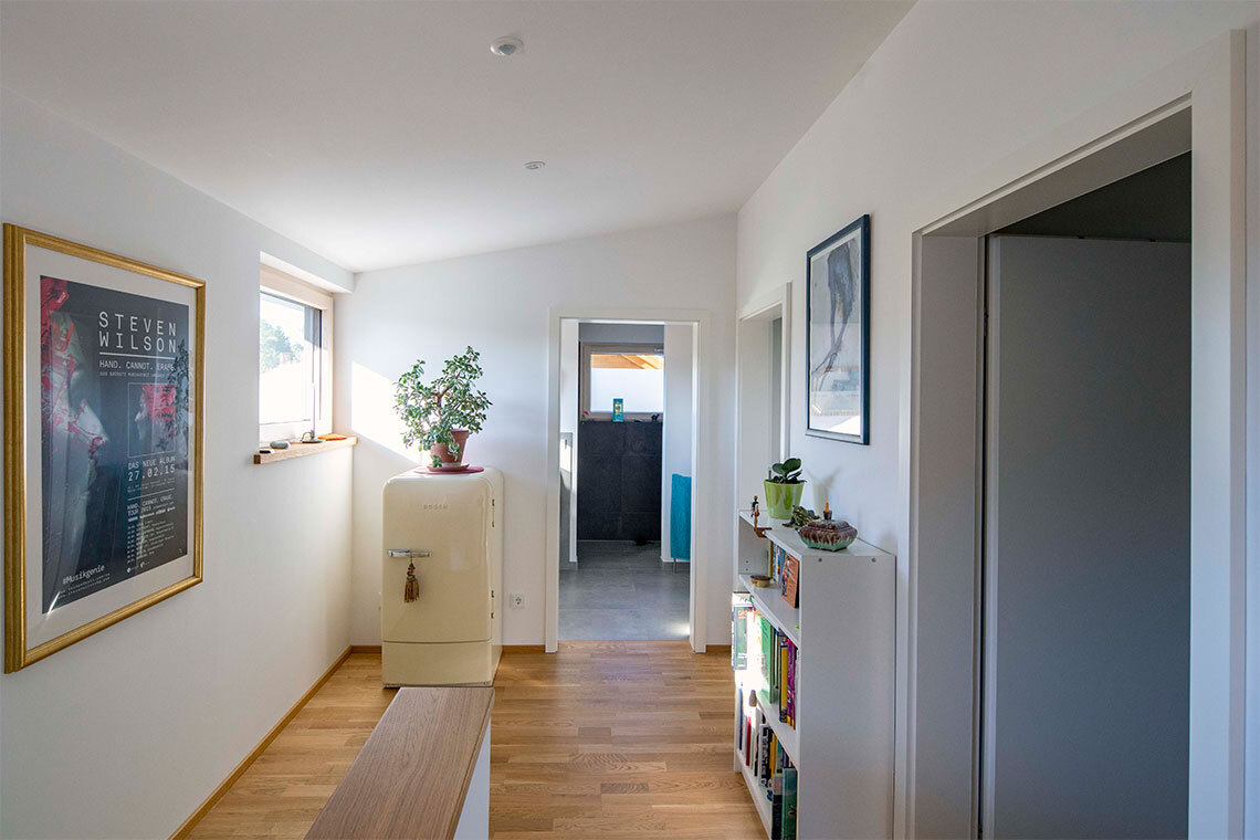 Ein heller Flur im Passivhaus Bad Aibling zeigt Pflanzen, Kunstwerke und einen cremefarbenen Kühlschrank vor einem Fenster.