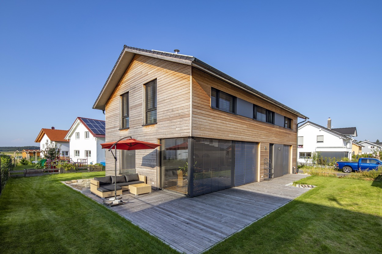 Ein modernes Holzhaus in Oberpframmern mit Terrasse und rotem Sonnenschirm steht in einer gepflegten Nachbarschaft.