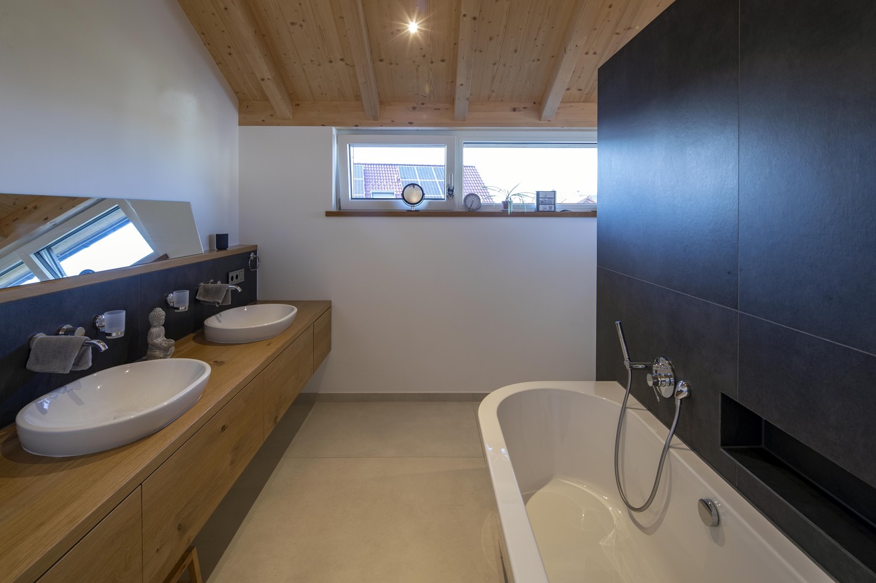 Modernes Badezimmer in einem Holzhaus in Oberpframmern mit freistehender Badewanne, zwei Waschbecken und eleganter Holzdecke.