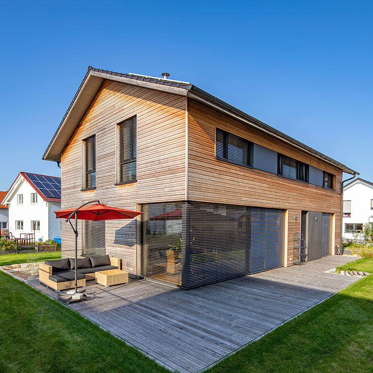 Modernes Holzhaus in Oberpframmern mit Terrasse, rotem Sonnenschirm und gepflegtem Rasen unter strahlend blauem Himmel.