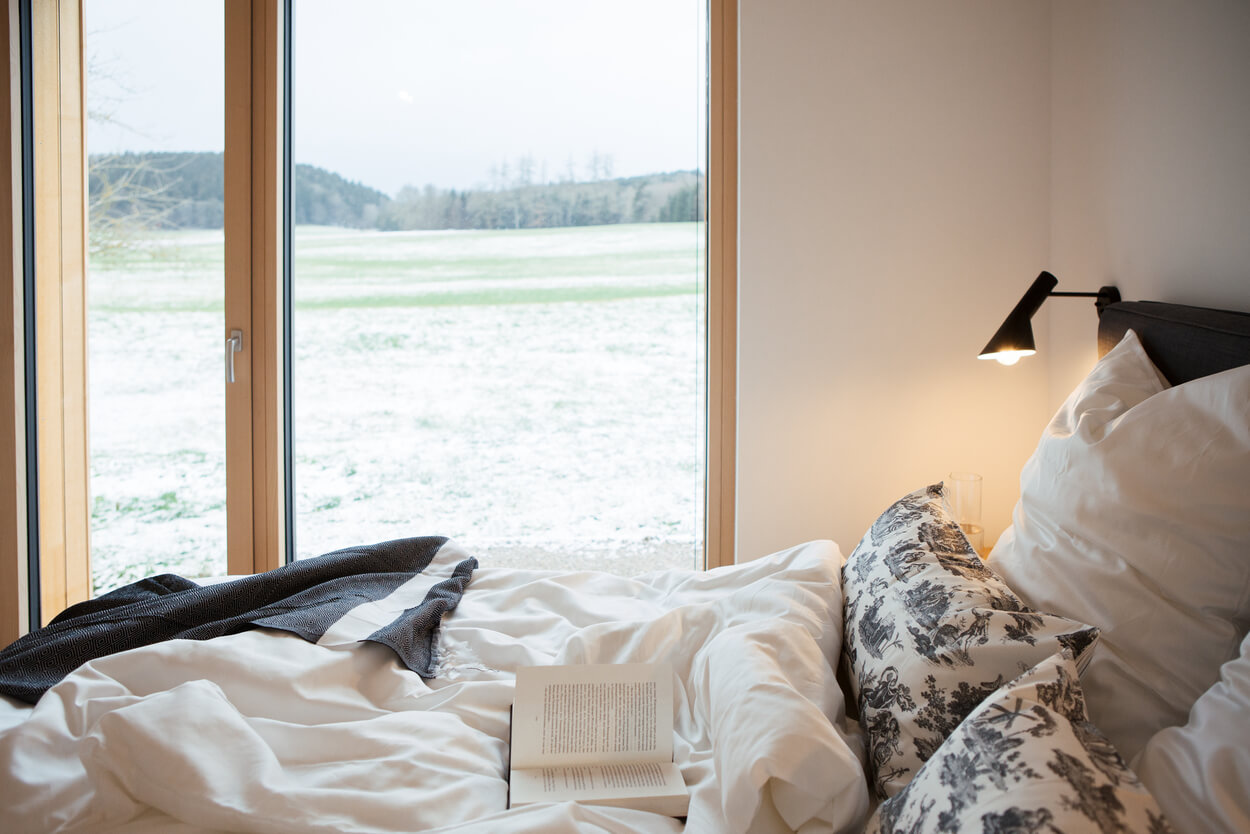 Ein gemütliches Bett mit offenem Buch lädt zum Entspannen im Holzhaus ein, während draußen der Chiemgau verschneit ist.