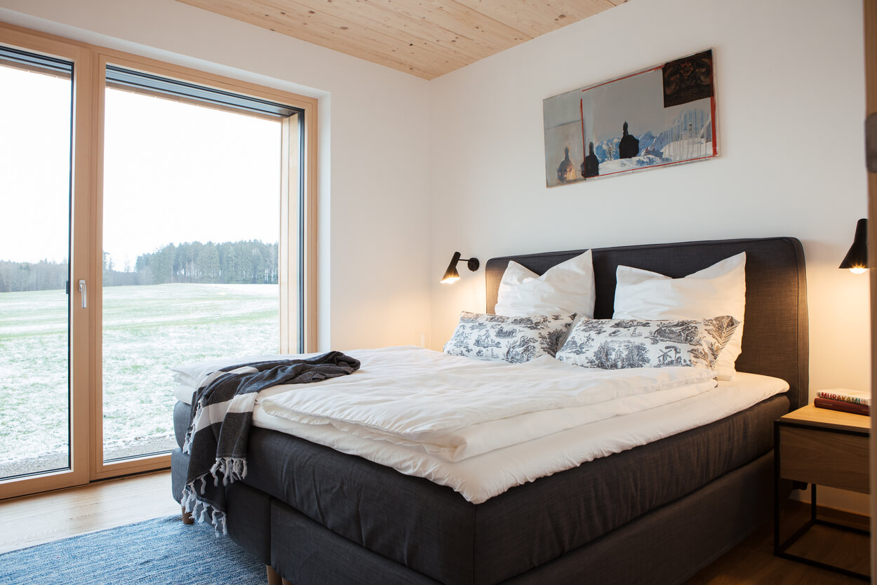 Ein gemütliches Schlafzimmer in einem Holzhaus im Chiemgau bietet durch ein großes Fenster einen Blick auf die schneebedeckte Landschaft.