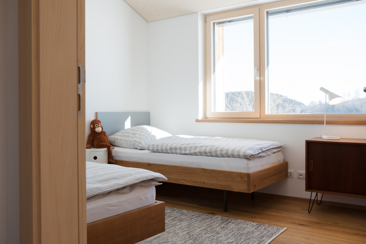 Ein helles Schlafzimmer in einem Holzhaus im Chiemgau mit zwei Betten, einem Plüschaffen und einem großen Fenster.