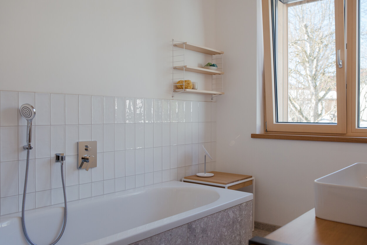 Ein helles Badezimmer im Holzhaus Chiemgau mit Badewanne, Duschkopf und großem Fenster bietet gemütliche Entspannung.
