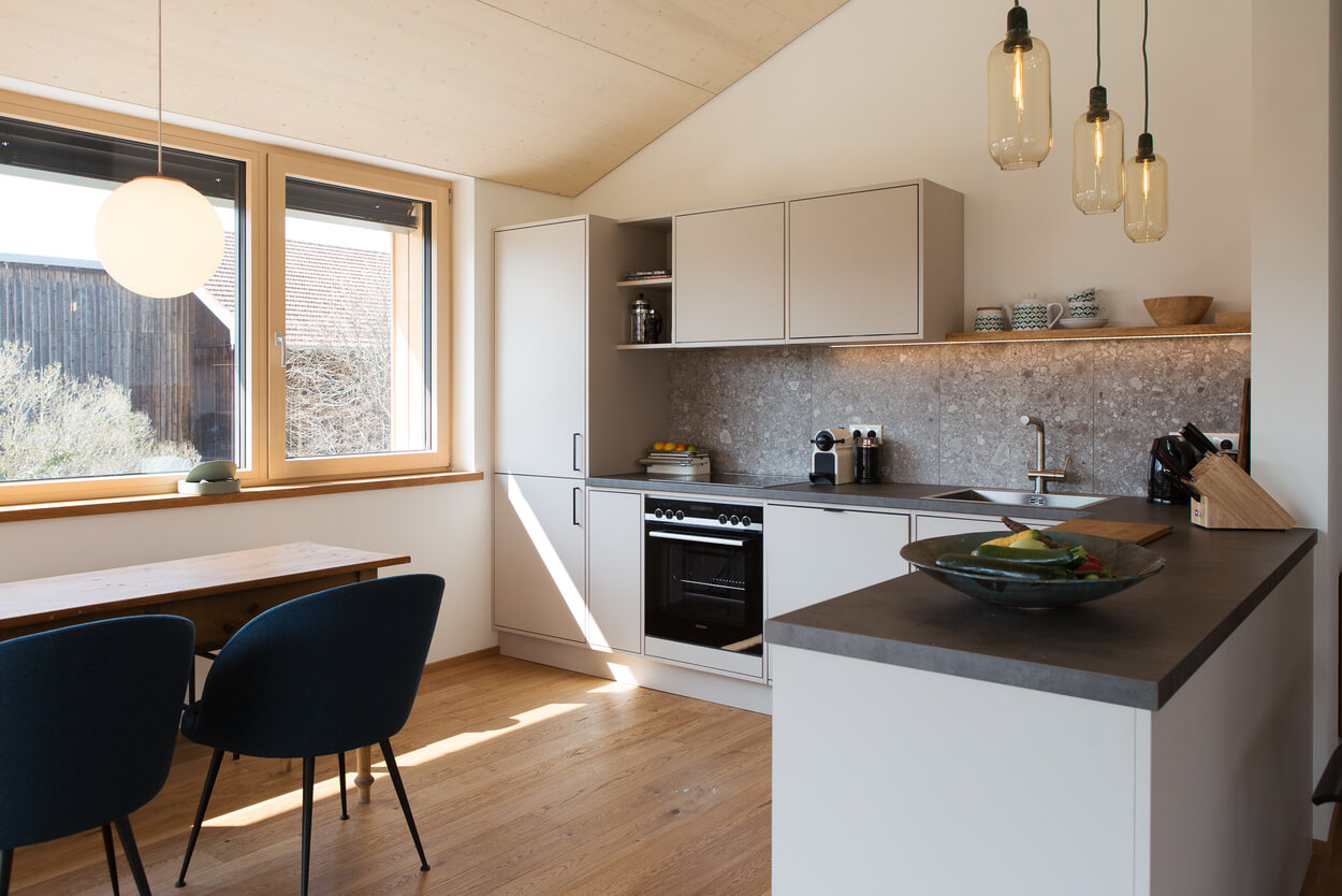 Moderne Küche im Chiemgau-Holzhaus mit großen Fenstern, stilvoller Beleuchtung und Holzboden strahlt Gemütlichkeit aus.