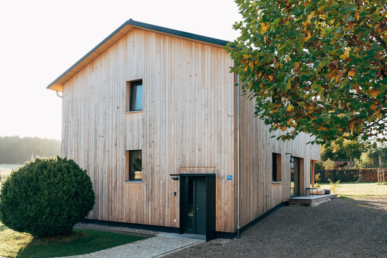 Ein modernes Holzhaus im Chiemgau steht idyllisch unter einem Baum in einer sonnigen, ländlichen Umgebung.
