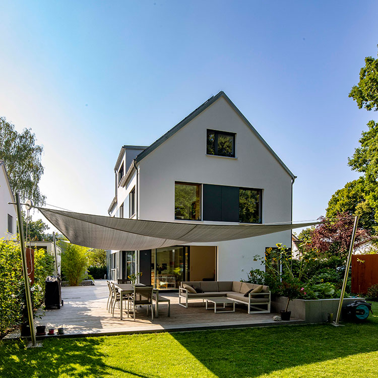Ein modernes Passivhaus in Eichenau mit Sonnensegel und gemütlicher Terrasse strahlt unter blauem Himmel.