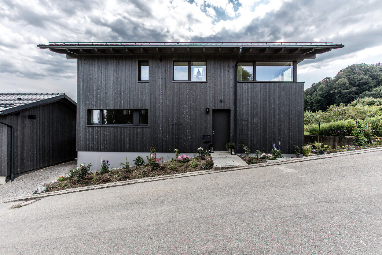 Ein modernes Holzhaus in Samerberg mit dunkler Fassade und gepflegtem Vorgarten steht vor einem bewölkten Himmel.