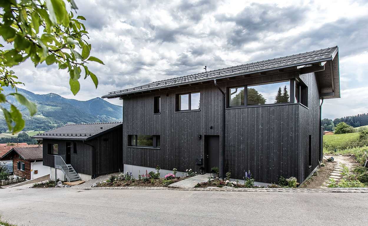 Ein modernes Holzhaus thront in Samerberg, umgeben von grünen Hügeln und einem bewölkten Himmel.