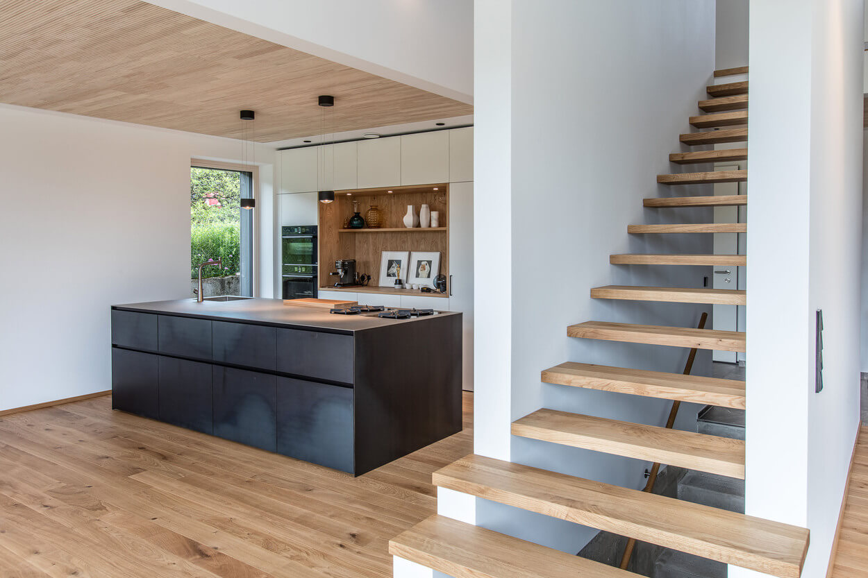 Die moderne Küche im Holzhaus Samerberg zeigt eine elegante Treppe neben einer stilvollen Kücheninsel mit Holzfußboden.