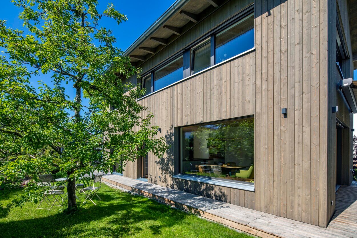 Modernes Holzhaus in Otterfing mit großer Fensterfront, umgeben von einem grünen Garten und einem schattenspendenden Baum.